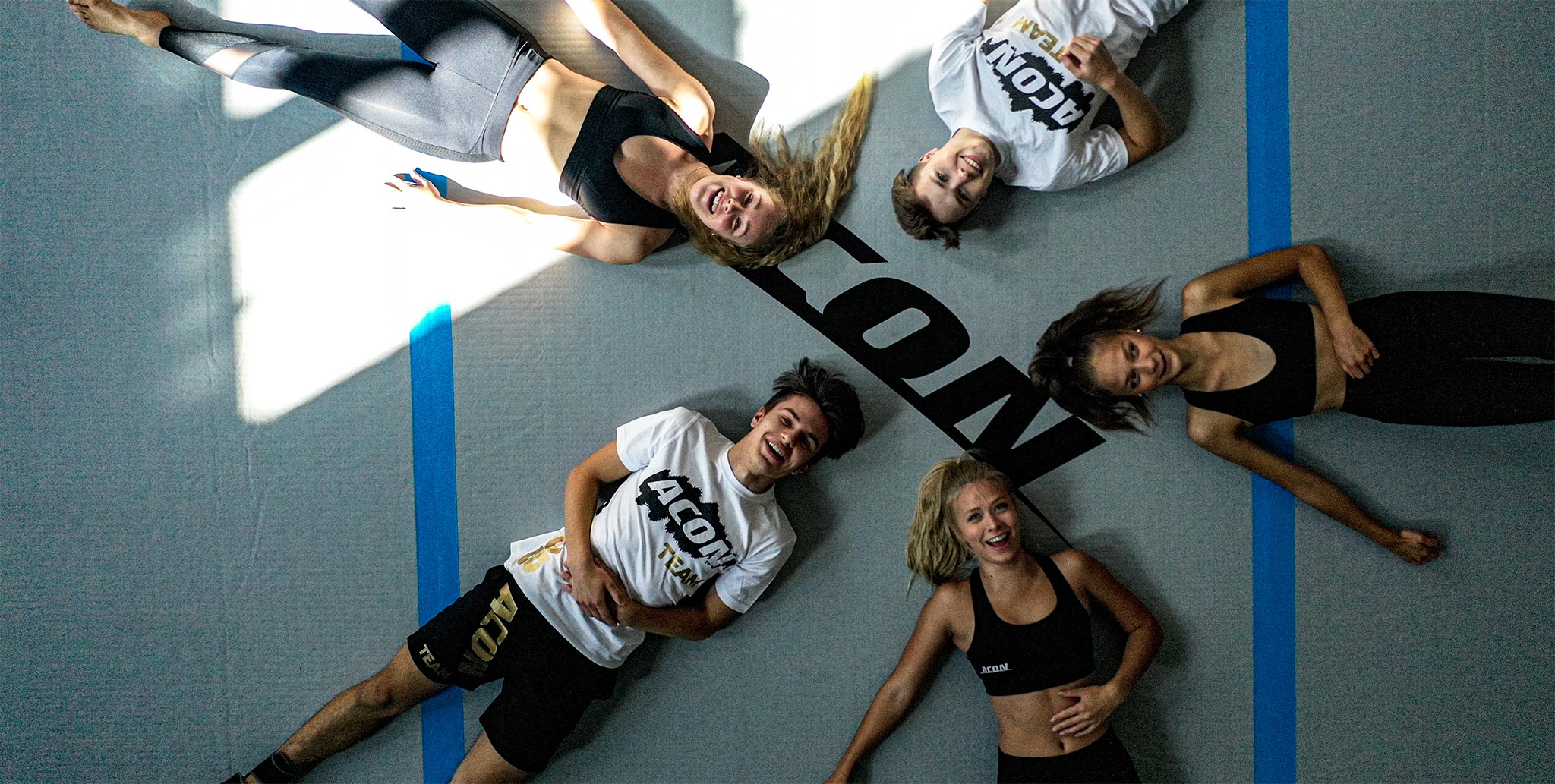 Cinq membres de l’équipe Acon, couchés sur un grand tapis Airtrack. L’image, prise de dessus, montre trois femmes et deux hommes. Ils sourient tous les cinq.