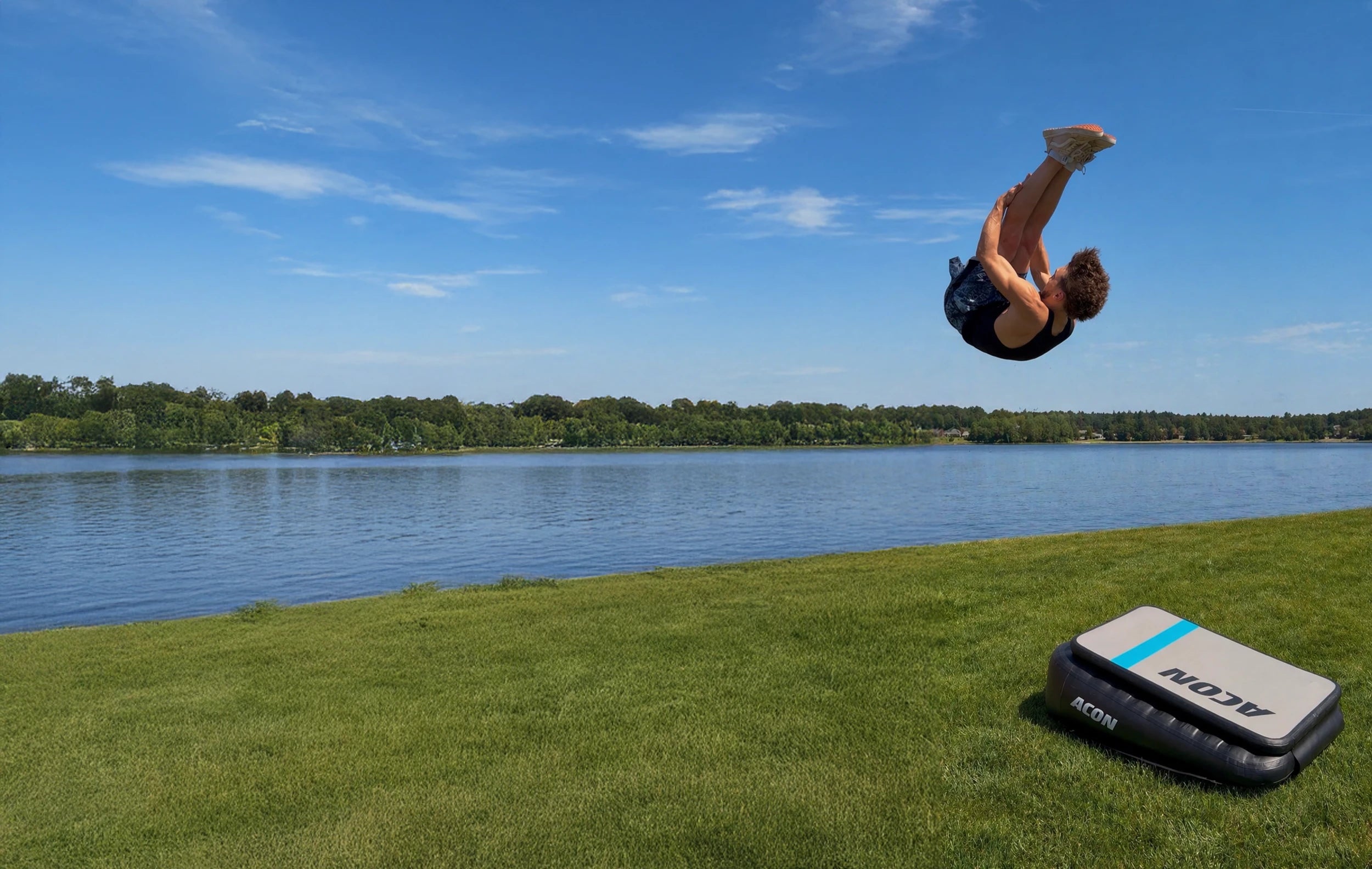 Un homme effectue un Pike Front sur un Acon Airboard au bord d’une rivière, l’Airboard posé sur l’herbe.