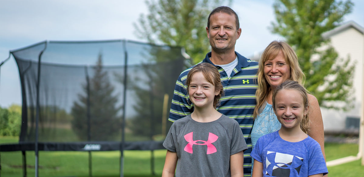 Une famille de clients Acon, composée d’un père, d’une mère et de deux filles. Il y a un trampoline ACON rond avec filet en arrière-plan