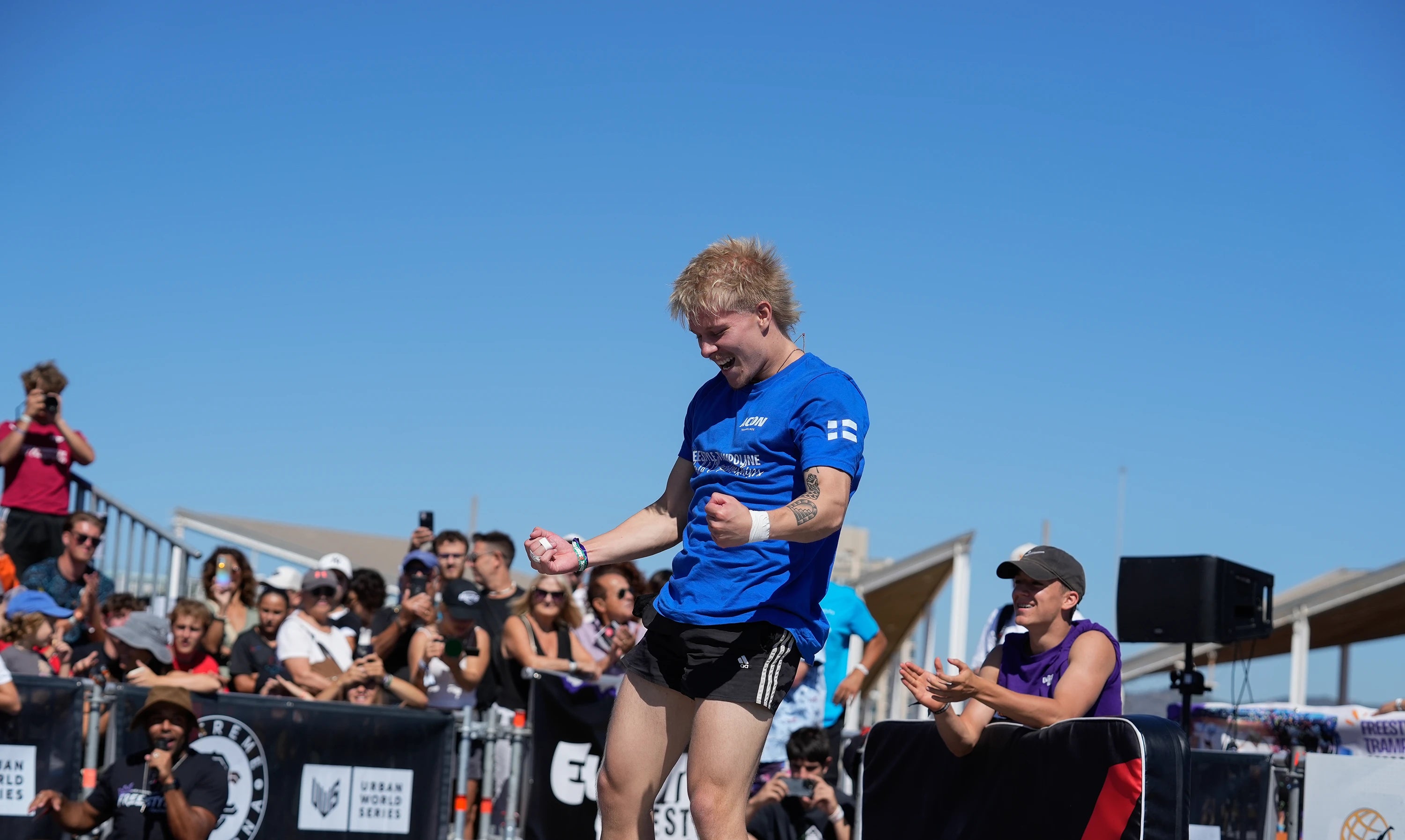 Homme en t‑shirt bleu et short noir sur un trampoline, avec des spectateurs en arrière-plan.