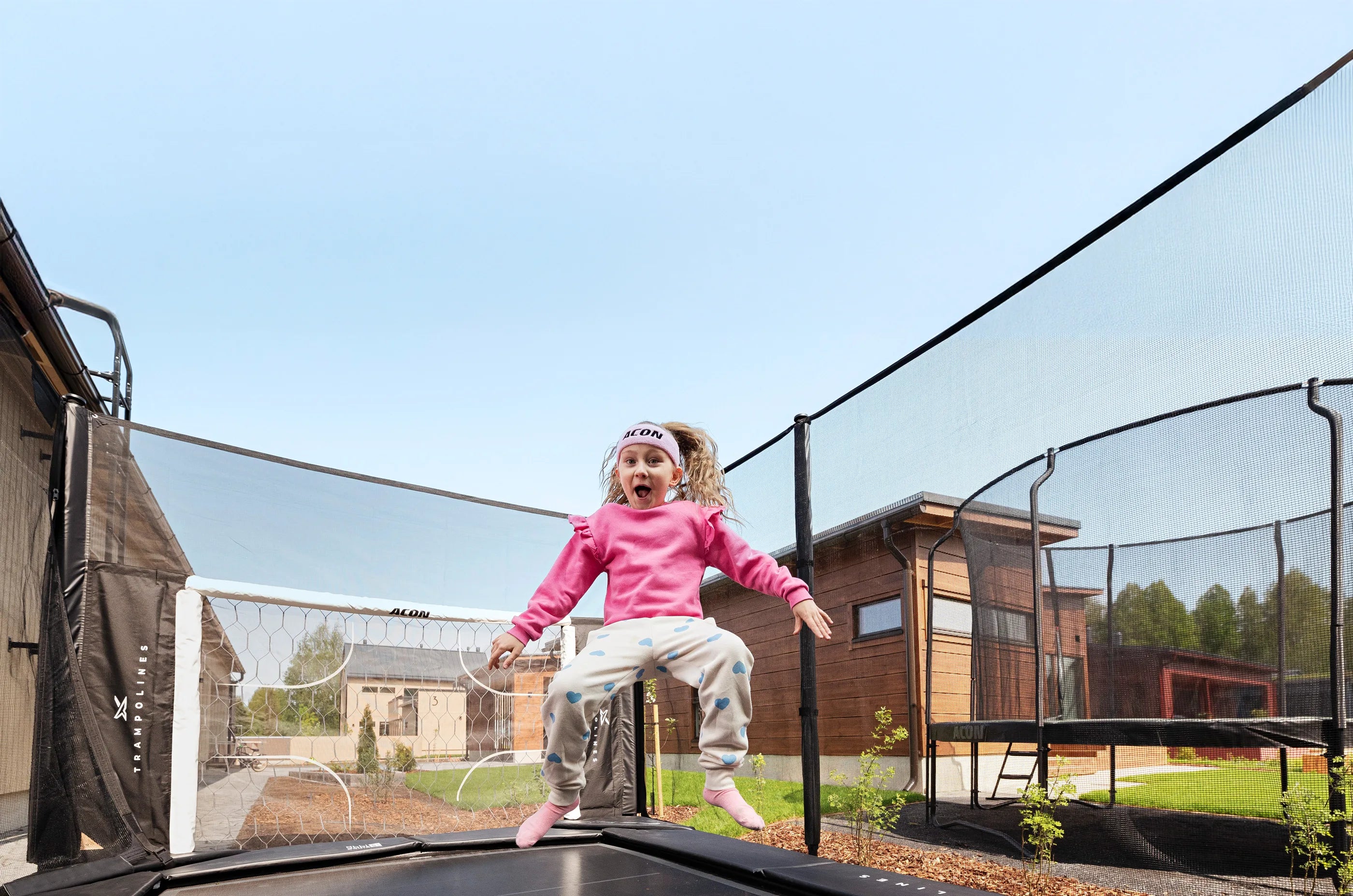 Une fillette joyeuse en t-shirt rose et bandeau Acon saute sur un trampoline Acon X, le sourire aux lèvres. Des maisons et un ciel bleu apparaissent en arrière‑plan.