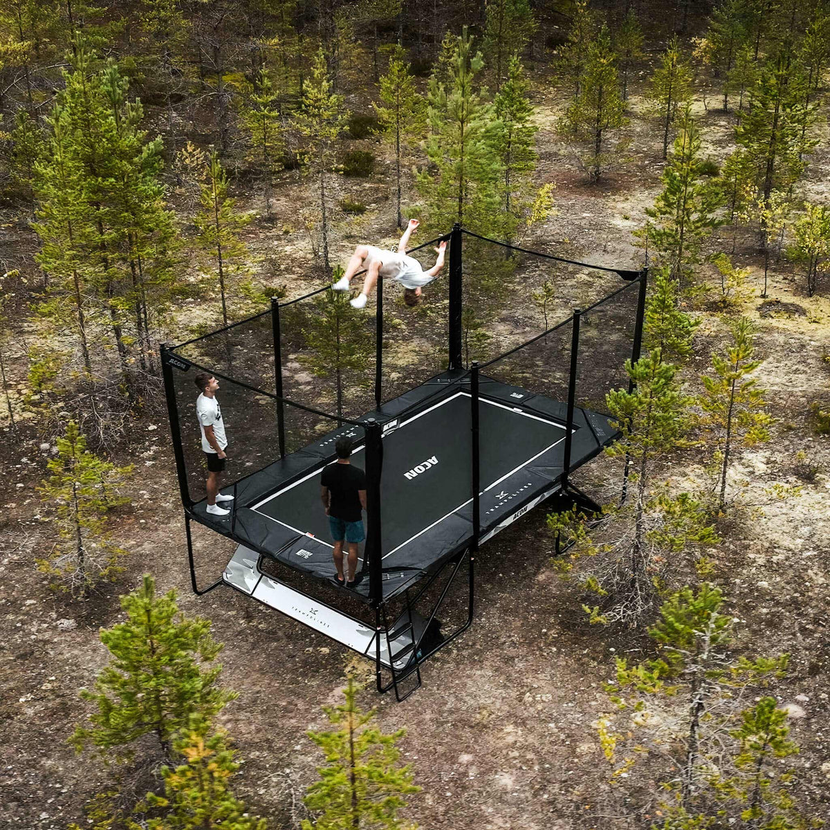 Trois trampolinistes sur un trampoline Acon X dans une forêt de pins nordiques.