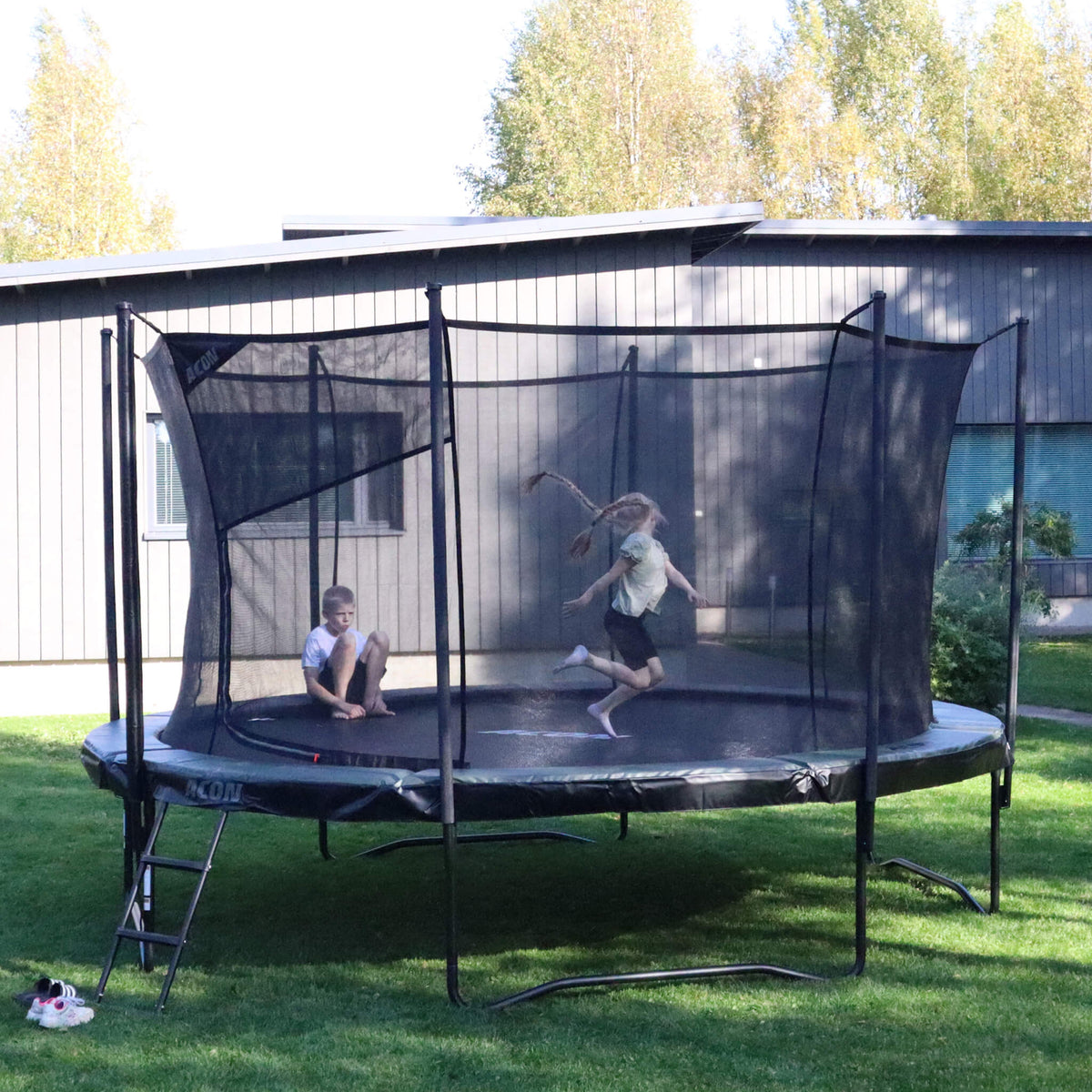 Trampoline Acon Air dans le jardin d’une maison individuelle. Un garçon est assis sur le trampoline, une fille saute.