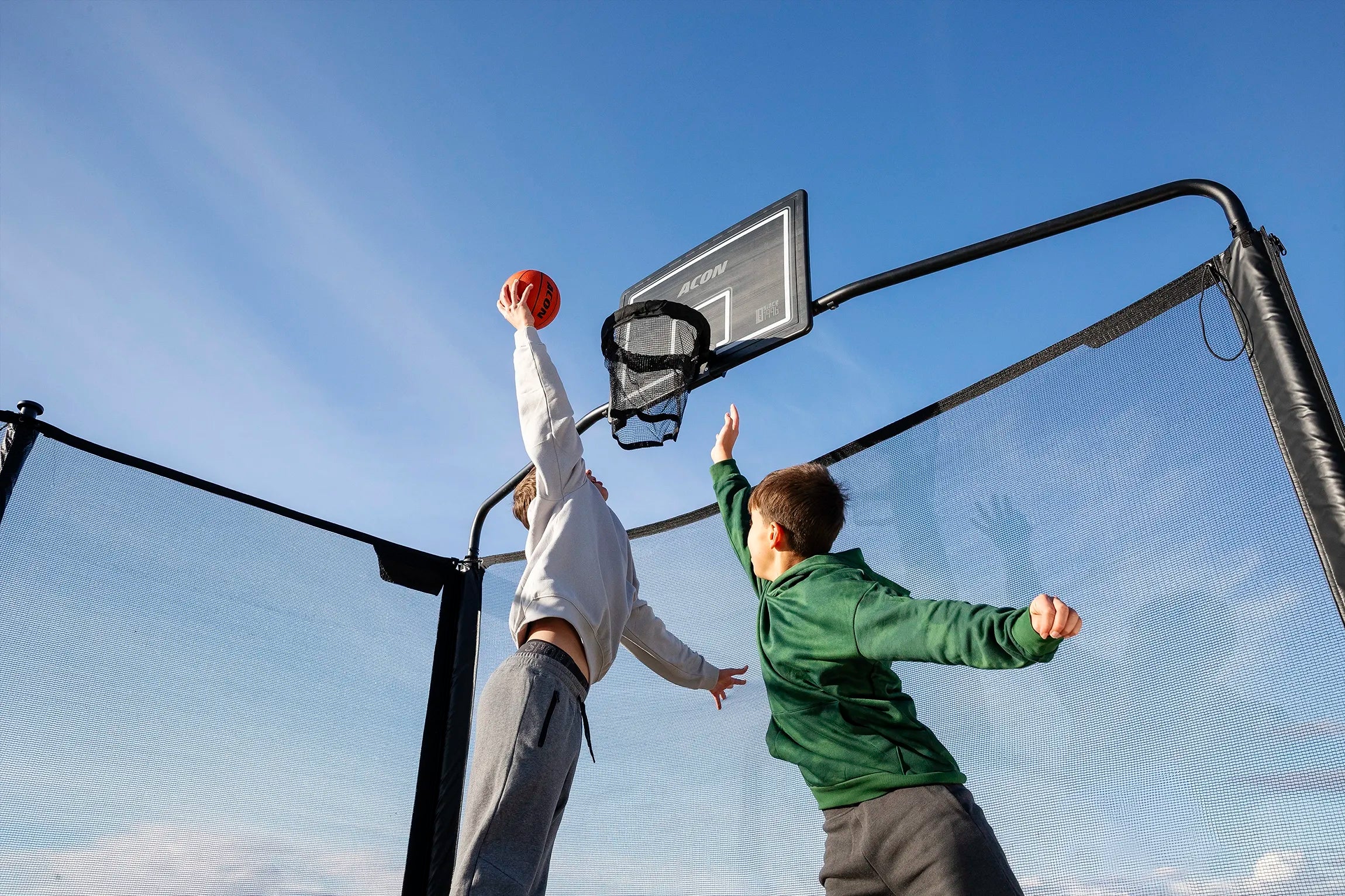 Deux personnes jouent au basket sur un trampoline Acon X en extérieur sous un ciel bleu clair.