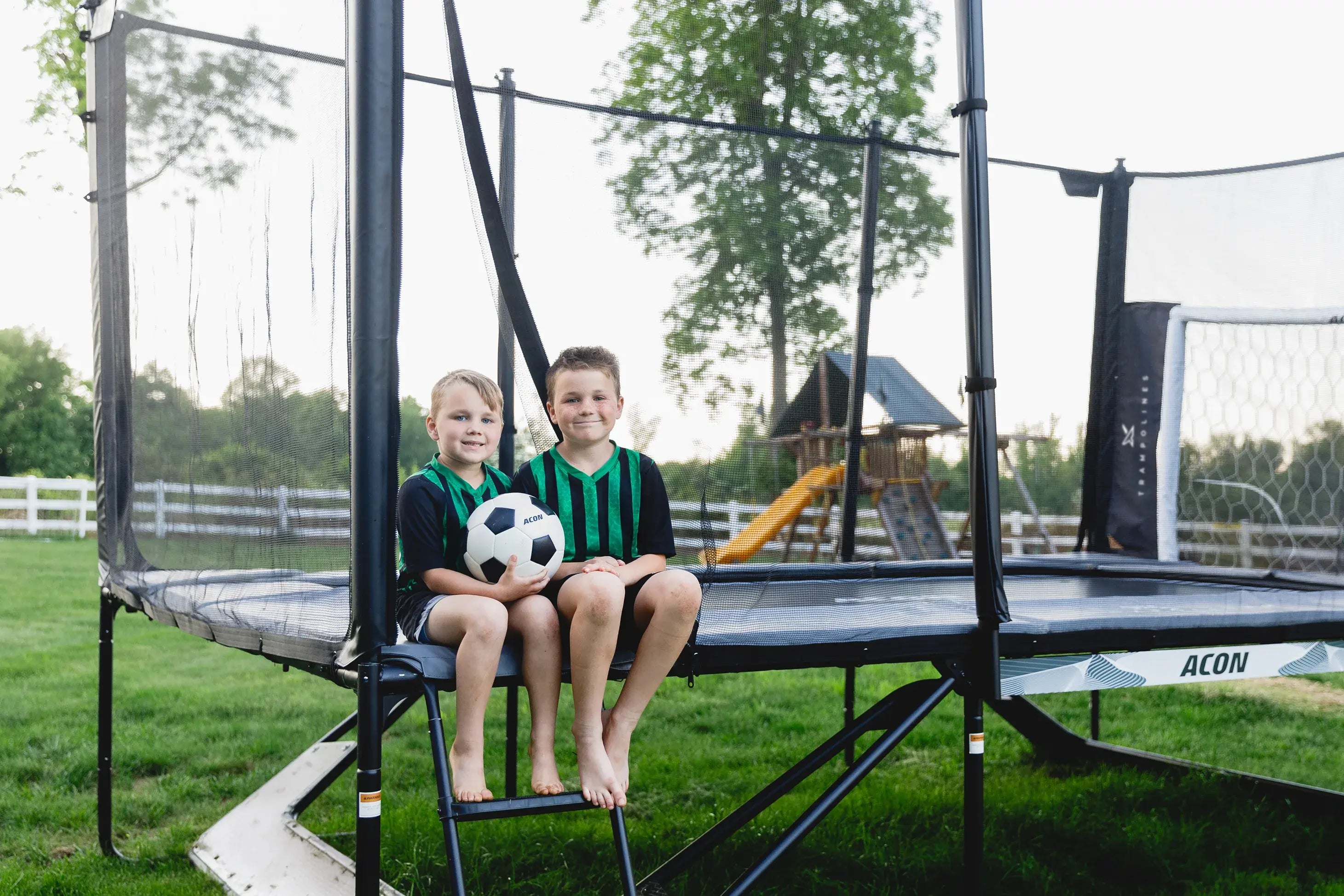 Deux enfants avec ballon de foot sur trampoline Acon dans le jardin avec filet et Panneau de but de football.
