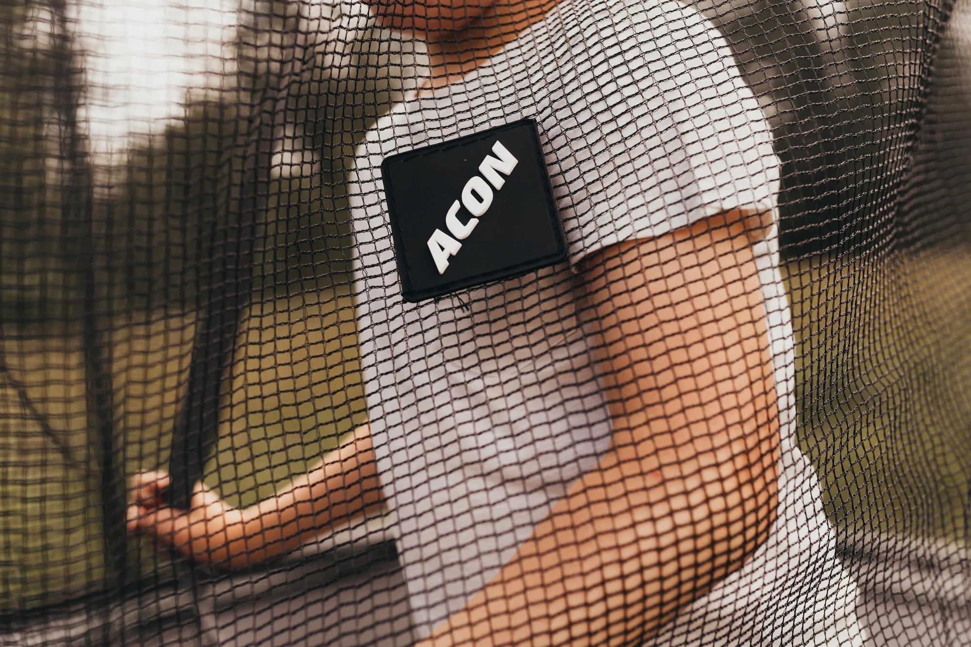 Un garçon portant un t-shirt blanc se tient derrière le filet de sécurité du trampoline sur lequel se trouve un logo ACON 