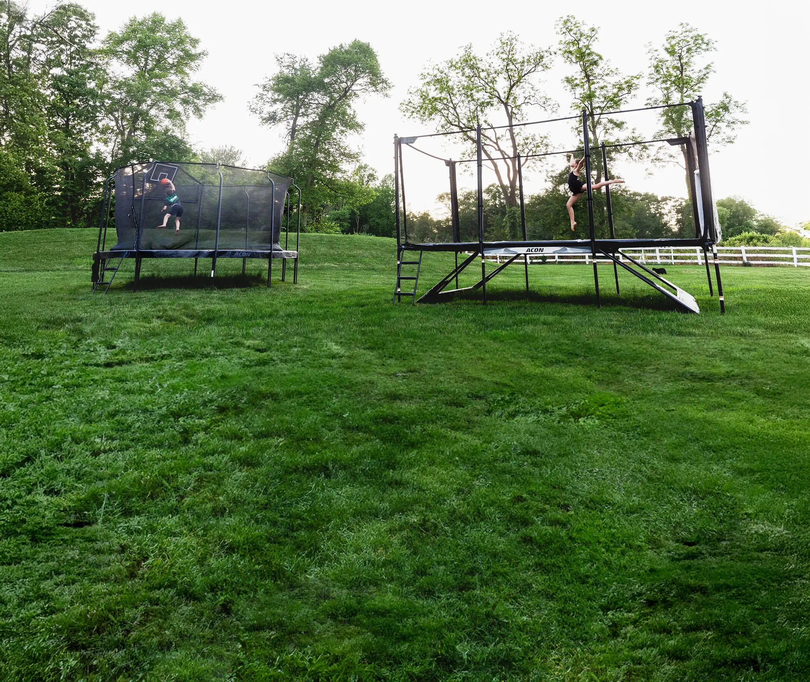 Un garçon joue au basket sur un trampoline Acon Premium, une fille saute sur un trampoline Acon X dans un jardin estival.
