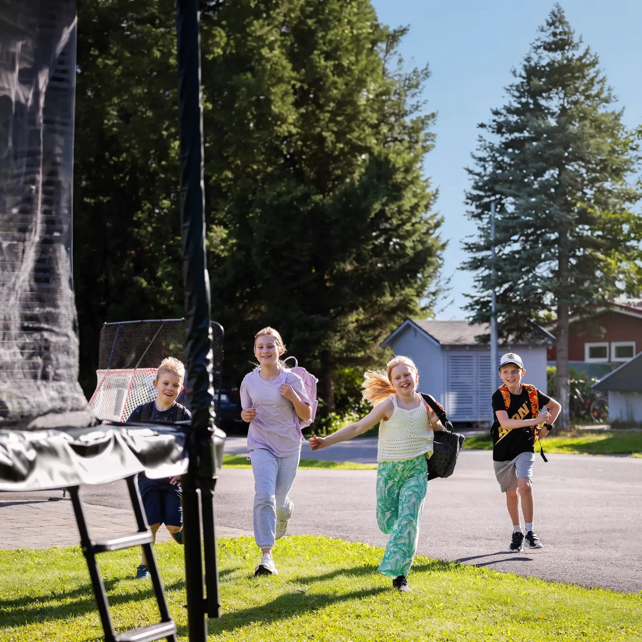 Quatre enfants courent vers un trampoline Acon après l’école, sacs au dos, dans un jardin ensoleillé.