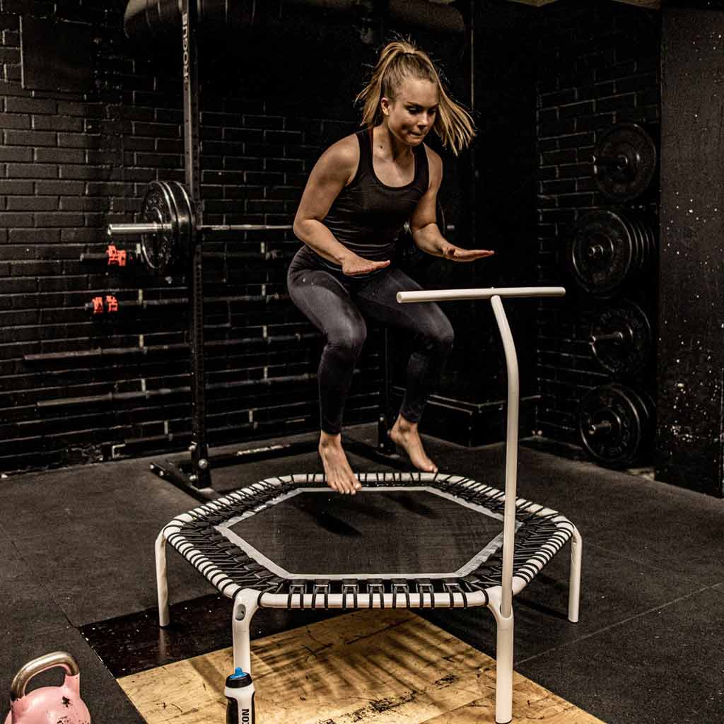 Une femme s'entraînant avec un trampoline hexagonal blanc ACON FIT avec guidon dans un gymnase intérieur