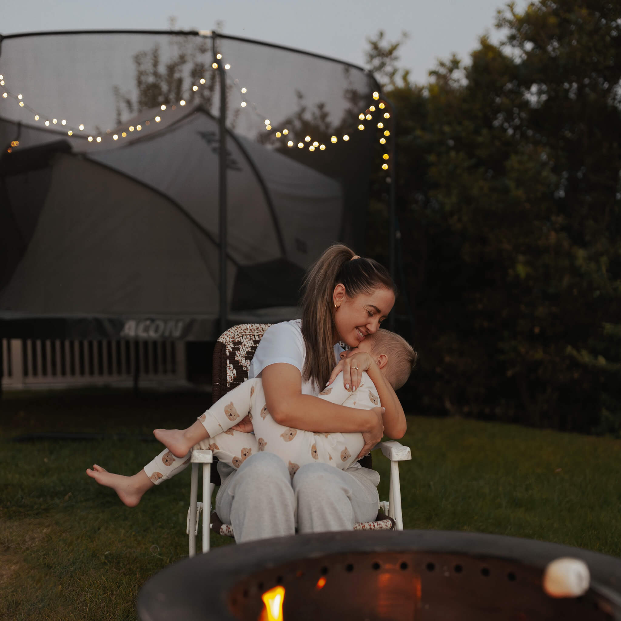 Mère étreignant son enfant, trampoline Acon décoré de lumières en arrière-plan, une tente de trampoline Acon dessus.