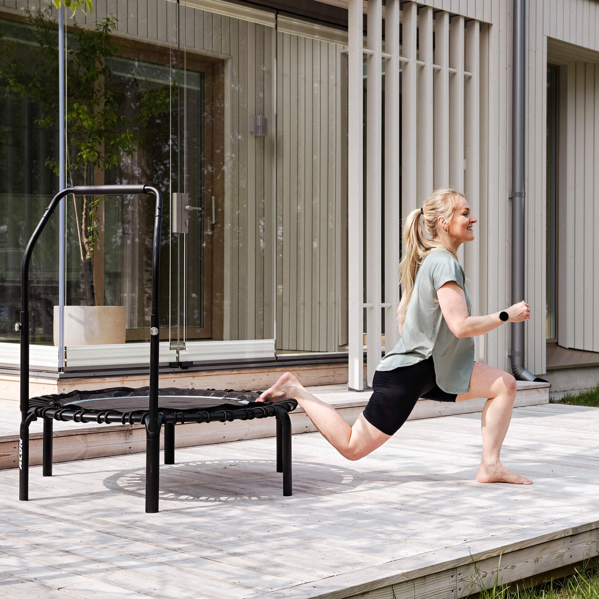Une femme fait des exercices de flexion sur un rebounder Acon FIT Balance sur la terrasse d’une maison.