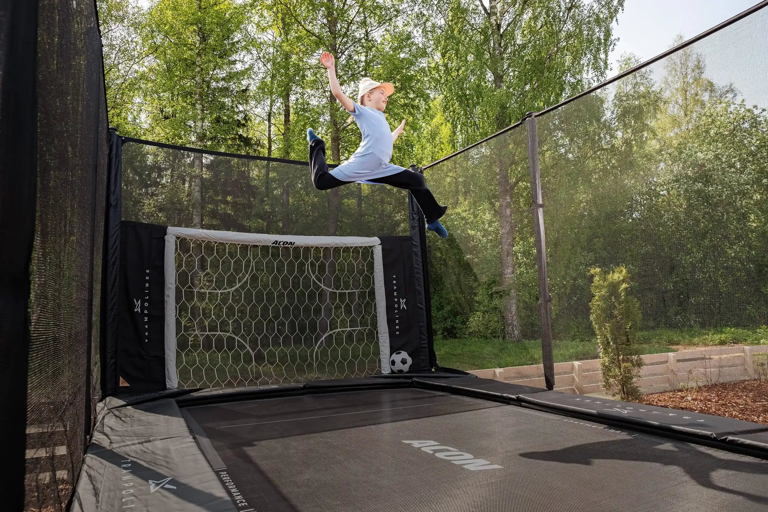 Fille sautant sur un trampoline Acon X dans un jardin estival, équipé d’un but de football Acon.