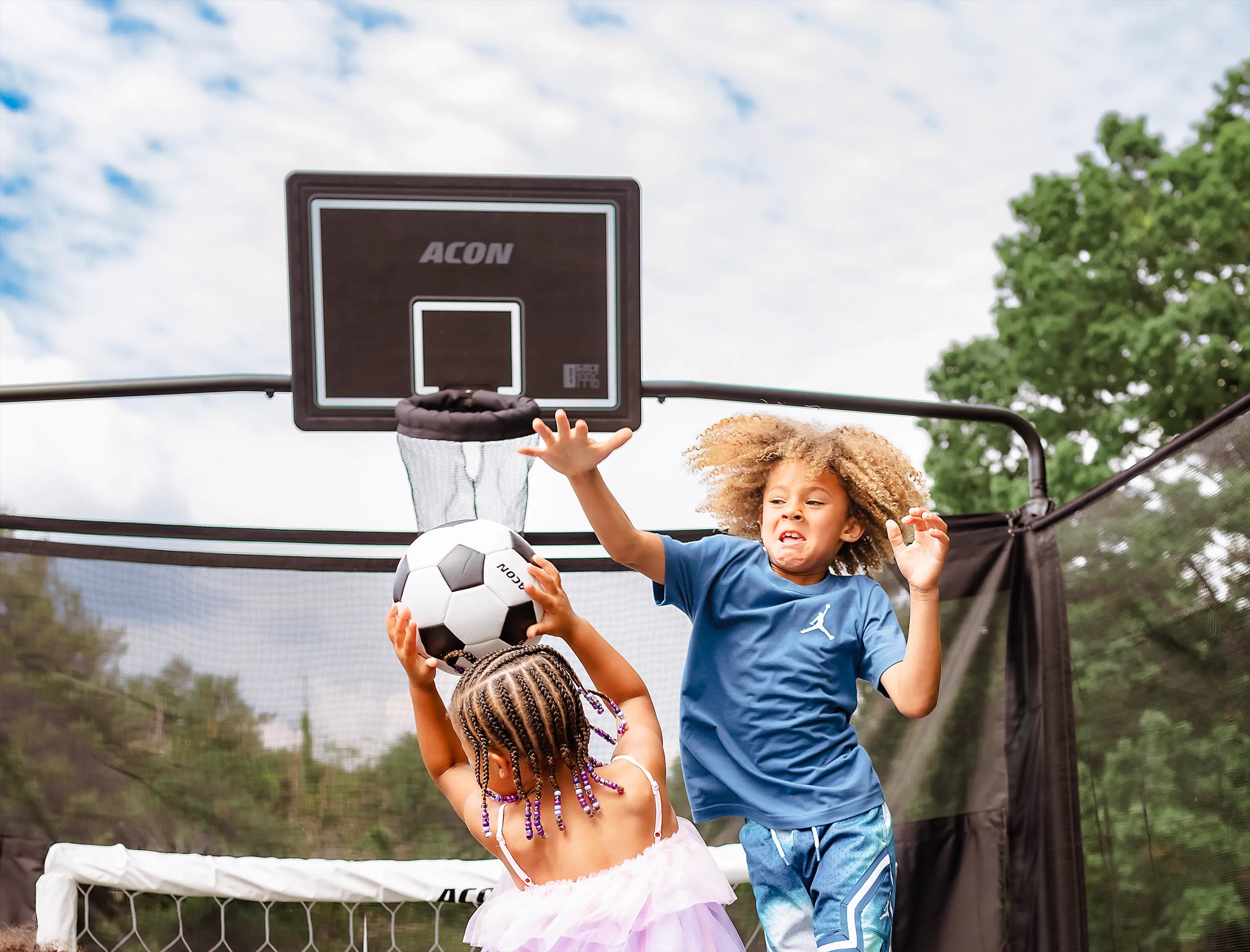 Deux enfants jouent au football sur un trampoline Acon X avec un but de football et un panier de basket.