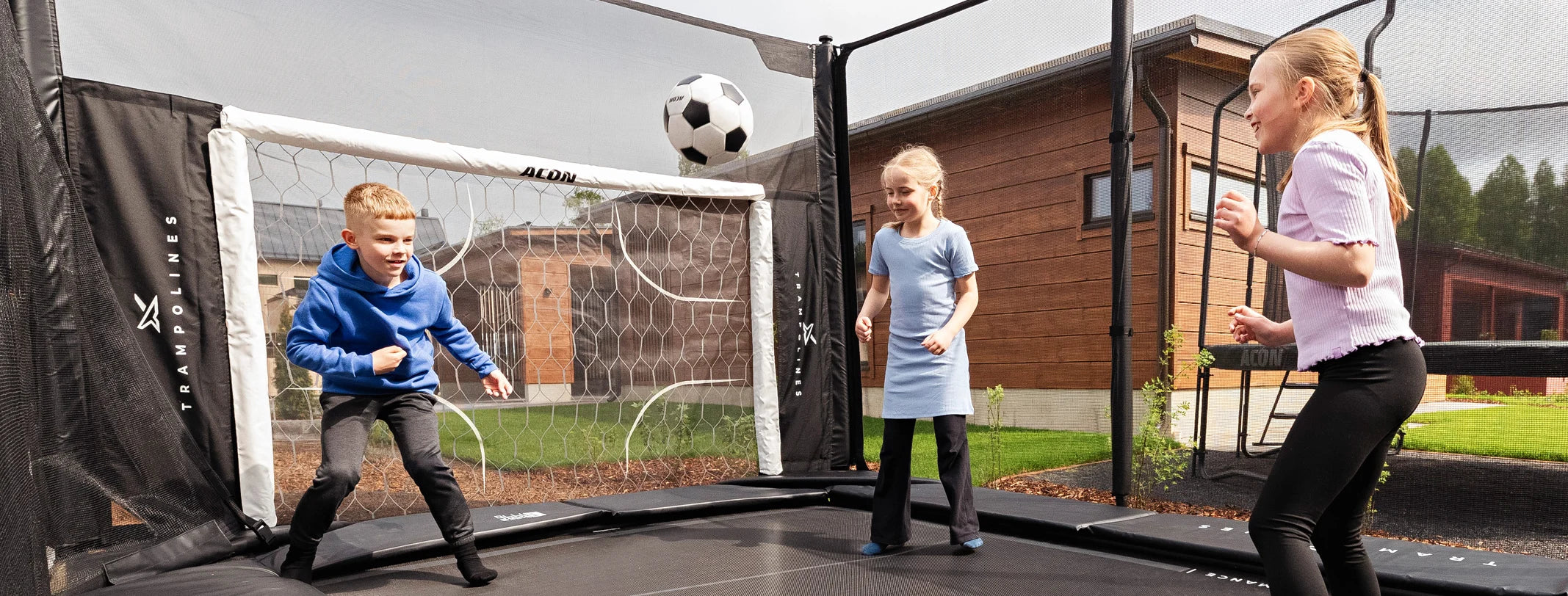 Trois enfants jouent au football sur un trampoline Acon X dans le jardin d'une maison individuelle. À une extrémité du trampoline se trouve l'accessoire But de football pour trampoline Acon X, vers lequel les enfants dirigent leurs tirs.