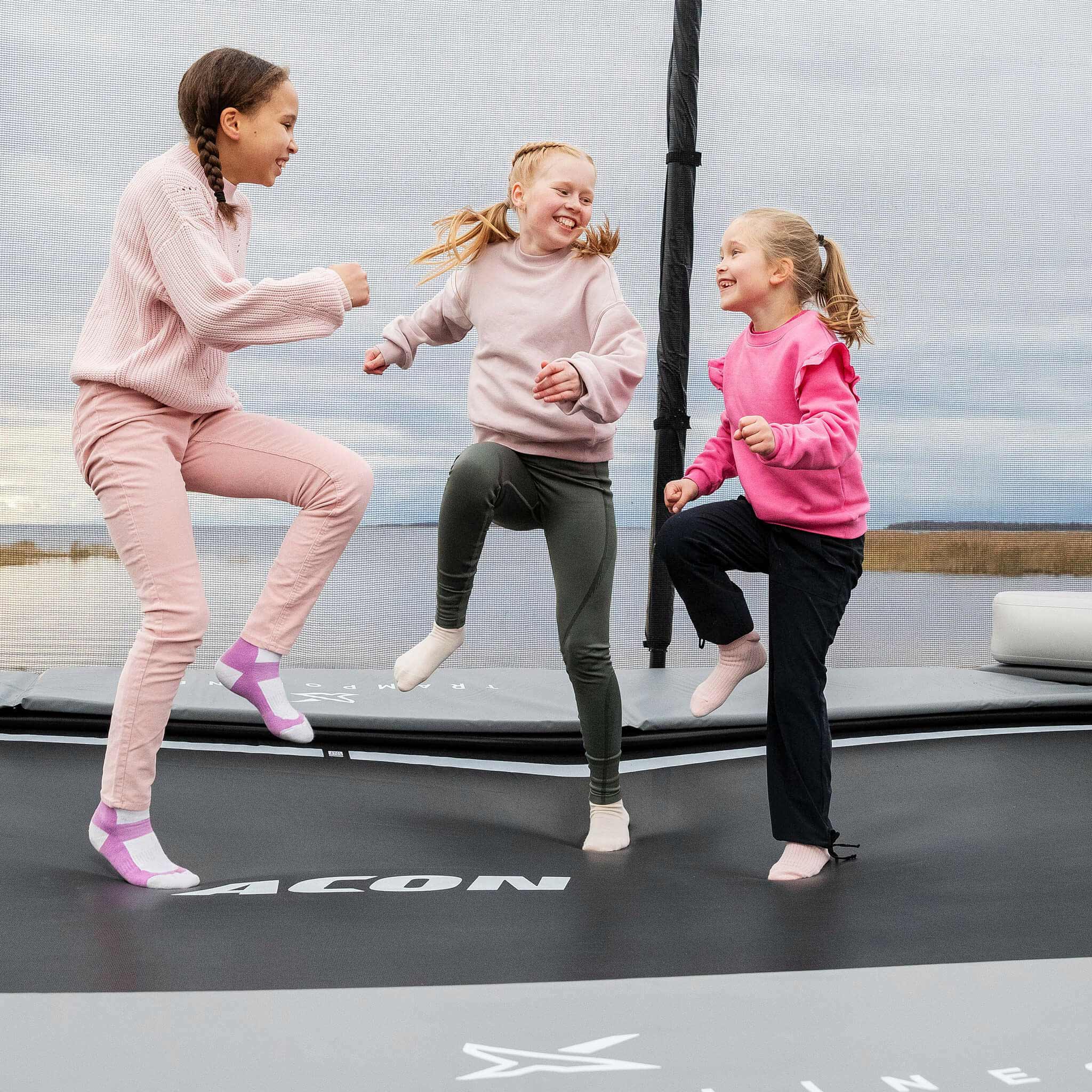 Trois filles joyeuses en chemise rose montent sur le trampoline Acon X. Une fille saute sur le trampoline Acon X. Deux filles se tiennent debout et regardent.