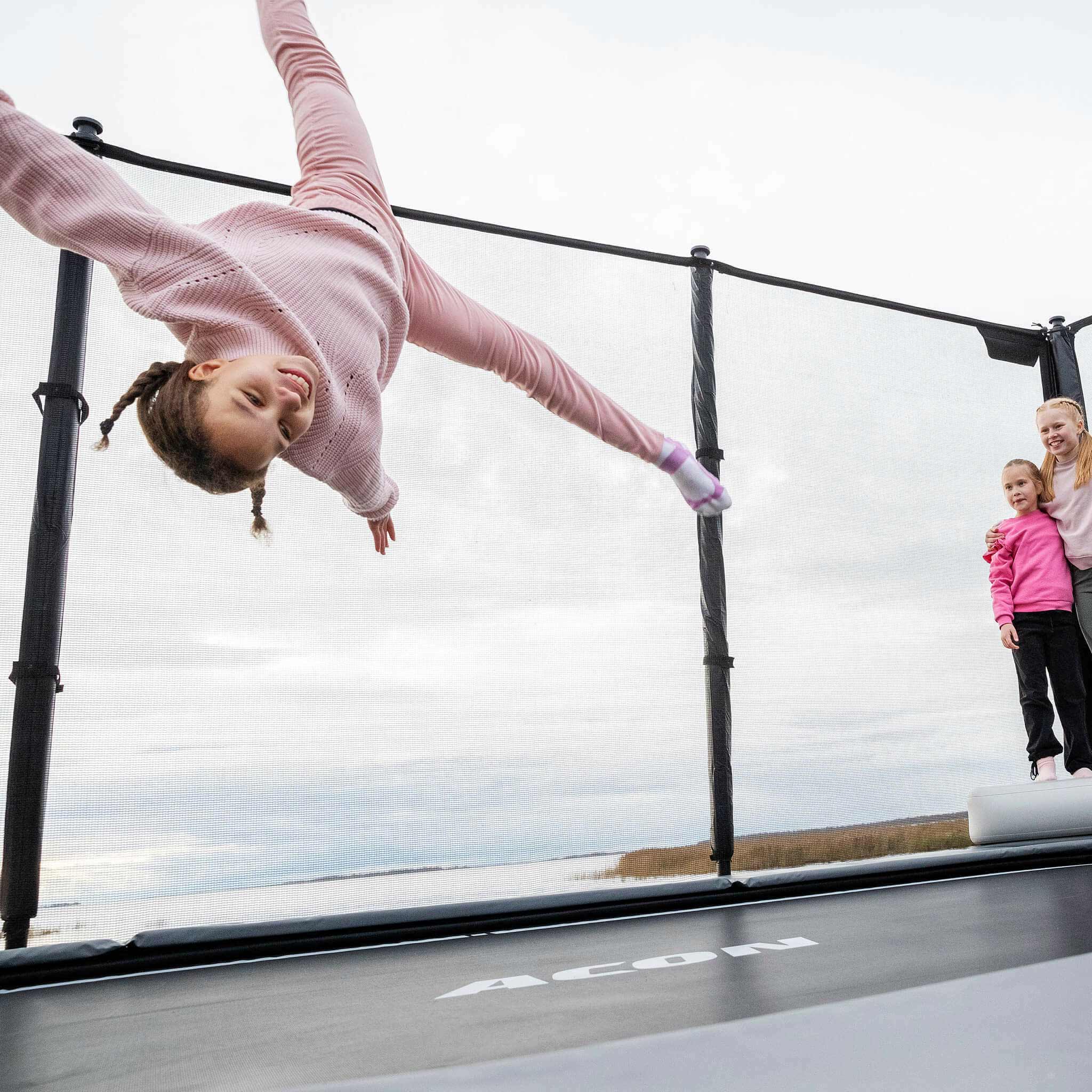 Une fille saute sur le trampoline Acon X, deux filles se tiennent debout et regardent.